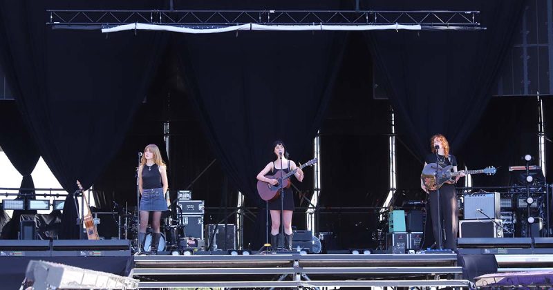 As tr&ecirc;s integrantes do grupo australiano Folk Bitch Trio tocam guitarras ac&uacute;sticas sob o sol de Lisboa, no MEO Kalorama 2026